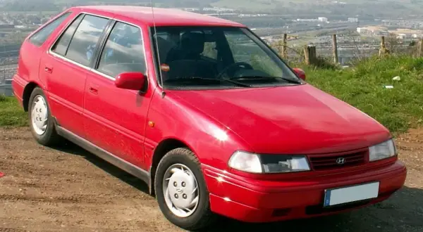 Czerwony Hyundai Pony z lat 90. stoi na brudnej drodze, z widokiem na dolinę i miasto w tle.