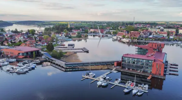 Widok na nowoczesny hotel i przystań jachtową na Mazurach. Widać liczne łodzie i żaglówki, a w tle miasteczko z charakterystyczną architekturą. To idealne mazury atrakcje dla miłośników żeglarstwa i wypoczynku nad wodą.