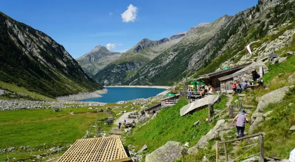 Ein idyllisches Bergpanorama mit einem türkisfarbenen See, umgeben von steilen Hängen. Hütten und Wanderer beleben das Bild, das an ein kleines Tibet im Zillertal erinnert.