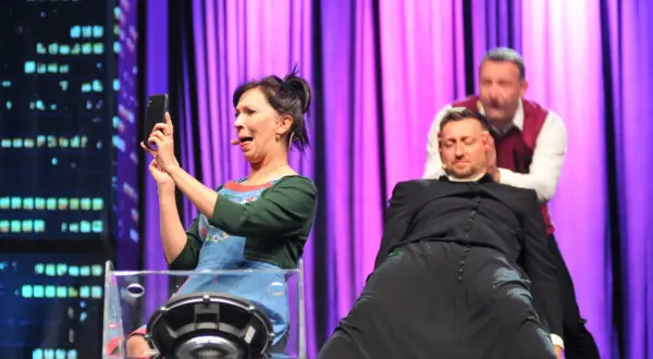 Trzech aktorów na scenie: kobieta robi selfie, mężczyzna w sutannie i drugi mężczyzna za nim.