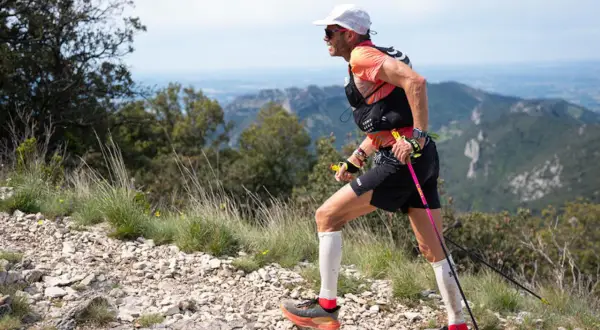 Uomo con bastoncini da trail running cos'è, corre su sentiero roccioso con vista panoramica su montagne e cielo azzurro.