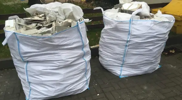 Dwa białe worki na gruz z niebieskimi szwami, gotowe do odbioru. W środku widać resztki materiałów budowlanych.