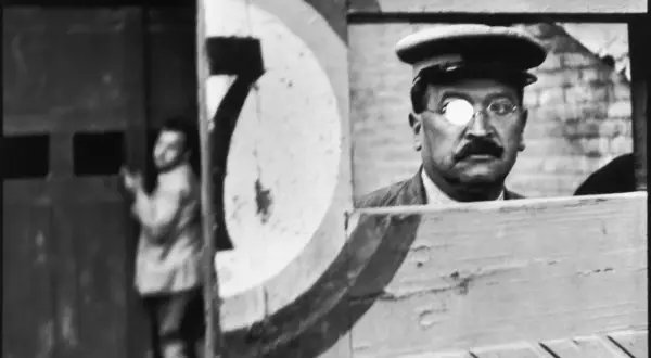 A man with a monocle and mustache, reminiscent of Cartier Bresson paintings, peers from behind a wooden structure. A blurred figure stands in the background.