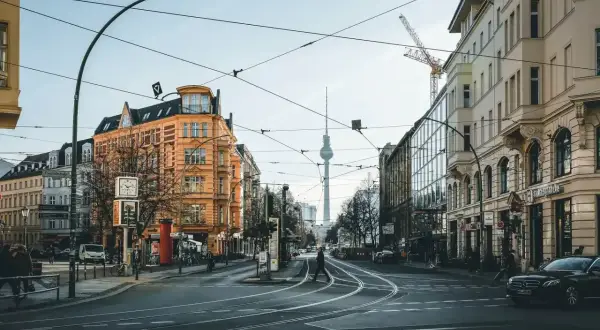 Skrzyżowanie w Berlinie z widoczną wieżą telewizyjną. Czy Berlin jest bezpieczny? Widać ludzi spacerujących, tramwaje i samochody.