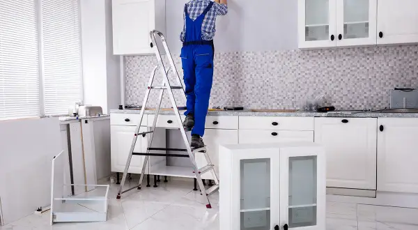 Un homme en salopette bleue est sur un escabeau, prêt à fixer un meuble au mur. La cuisine est en cours d'installation.
