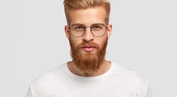 Un homme au visage rectangulaire, barbe rousse et lunettes rondes, porte un t-shirt blanc.