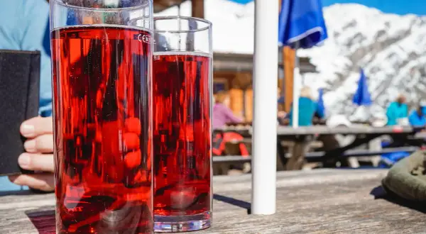 Zwei Gläser mit rotem Getränk, vielleicht ein Skiwasser-Rezept, stehen auf einem Holztisch. Im Hintergrund eine Skihütte mit Gästen.