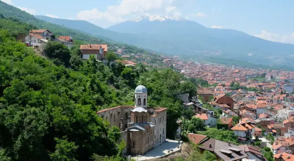 Eine malerische **Kosovo Landschaft** mit einer alten Kirche im Vordergrund, umgeben von üppigem Grün. Im Hintergrund erstreckt sich eine Stadt mit roten Dächern bis zu schneebedeckten Bergen.