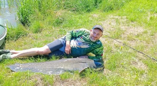 Szczęśliwy wędkarz z potężnym sumem złowionym w zbiorniku Nielisz. Obok łódka i wędka.