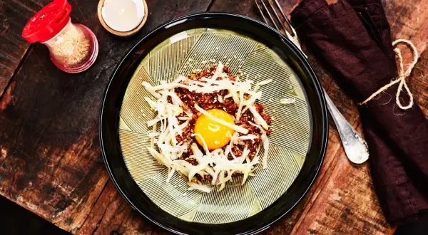 En läcker koreansk råbiff med en glänsande äggula i mitten, toppad med strimlad rättika och sesamfrön. Serveras på en vacker tallrik med en mörk servett och gaffel.