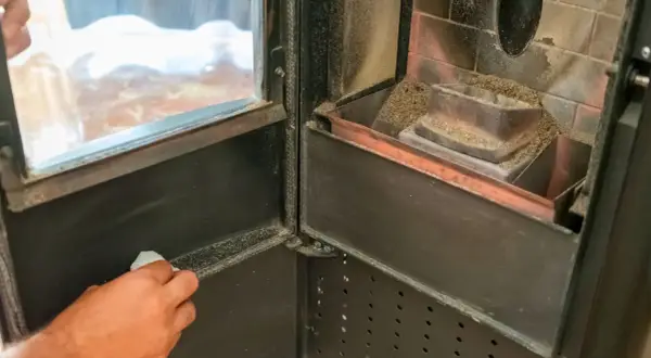 Mano che pulisce il vetro di una stufa a pellet, rimuovendo la cenere per una migliore visione del fuoco.