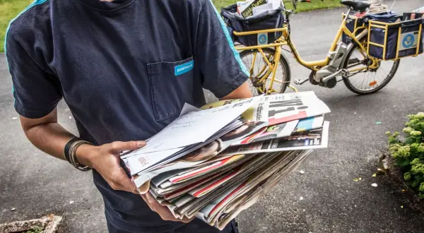 Postnord-utdelare med post i händerna, redo för postutdelning varannan dag. En gul postcykel syns i bakgrunden.