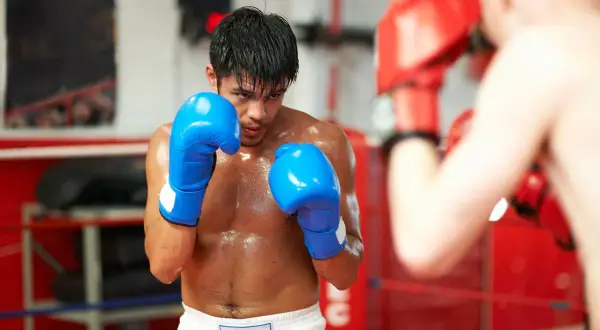 A boxer in blue gloves sweats during a tough boxing cardio workout in the ring, facing an opponent in red gloves.
