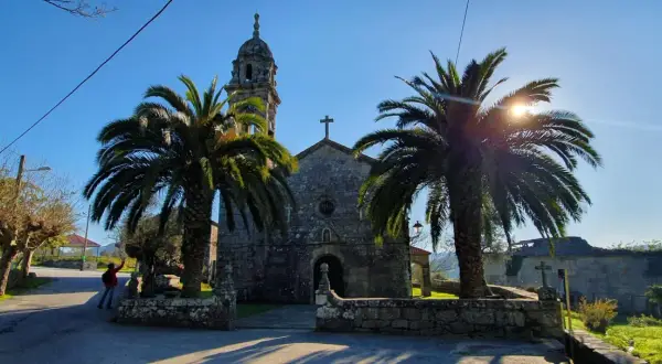 Iglesia de piedra con palmeras y sol brillante. Un lugar tranquilo que ver en Rianxo, ideal para pasear.