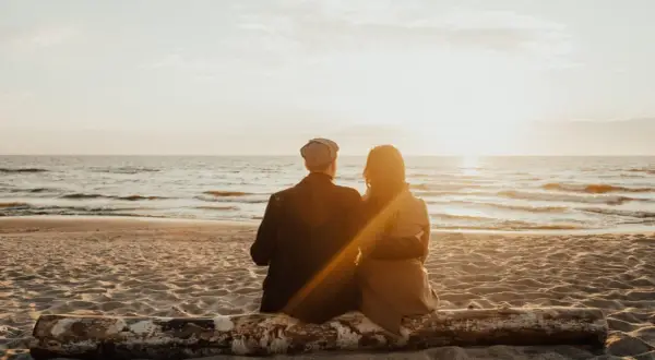 Para dwojga siedząca na pniu na plaży, patrząca na zachód słońca nad morzem. Idealne, fajne miejsce nad morzem dla dwojga.