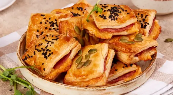 Pizzerinki z ciasta francuskiego, posypane czarnuszką i pestkami dyni, ułożone na talerzu. Idealne na przekąskę.
