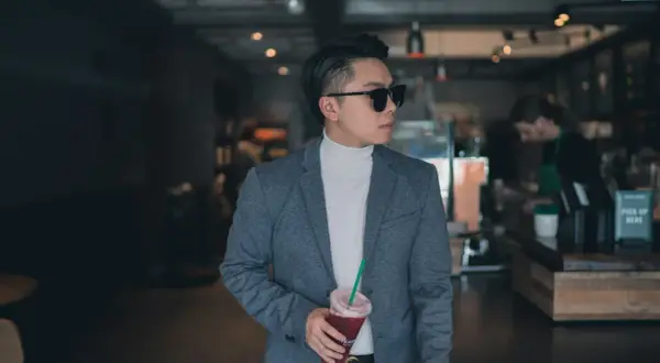 Mann im grauen Anzug mit weißem Rollkragenpullover und Sonnenbrille, hält Getränk. Das ist smart casual.