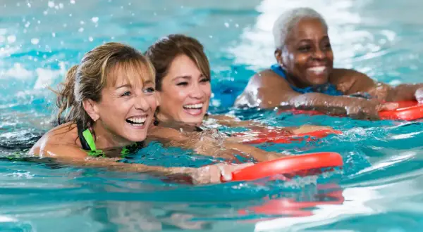 Three women joyfully participate in an adult learn to swim class, holding red kickboards as they move through the water.