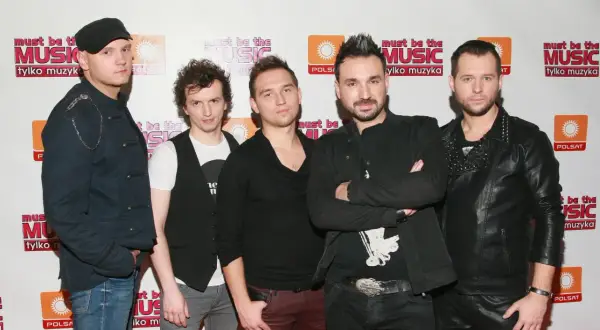 Pięciu mężczyzn w strojach scenicznych pozuje do zdjęcia na tle ścianki z logo "Must Be The Music".