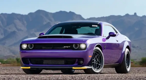 En lila amerikansk sportbil, en Dodge Challenger SRT, står parkerad på en grusväg med berg i bakgrunden.