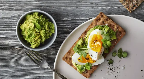 Delicioso sandwich de aguacate y huevo sobre pan integral, con un tazón de aguacate machacado y un tenedor al lado.