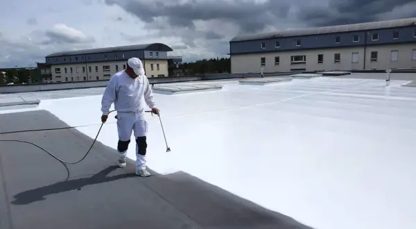Un ouvrier applique une peinture réflective toiture blanche sur un toit plat. Le ciel est couvert de nuages sombres.