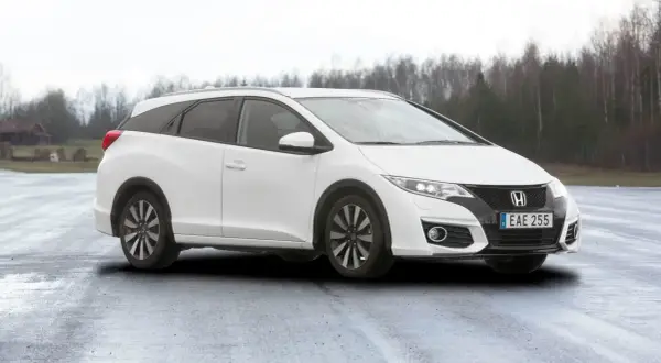 Vit Honda Civic Tourer parkerad på en fuktig väg med träd i bakgrunden.