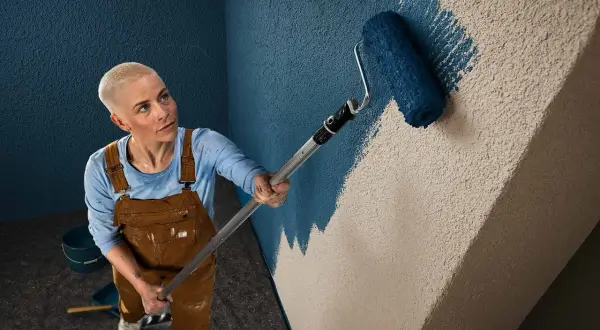 Frau mit Kurzhaarschnitt streicht mit einer Farbrolle eine Wand mit Rauputz. Sie trägt Latzhose und blaue Langarmshirt.