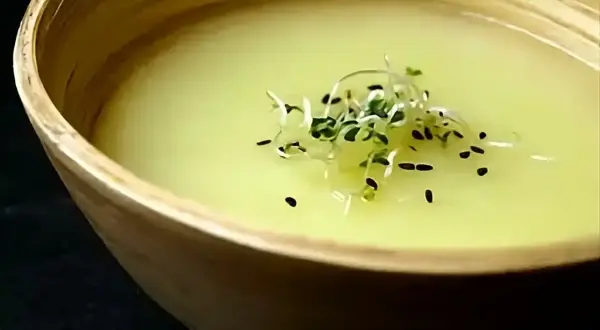 Un bol en bois contient un velouté d'asperges blanches, garni de pousses vertes et de graines noires.
