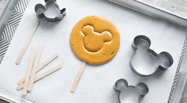 En färdiggjord dalgona cookie med Musse Pigg-form ligger på en bakplåt med flera Mickey Mouse-formar och träpinnar.