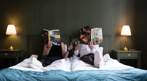 Två personer läser tidningar och skålar med champagne i en säng på Hotell Hertig Karl Filipstad.