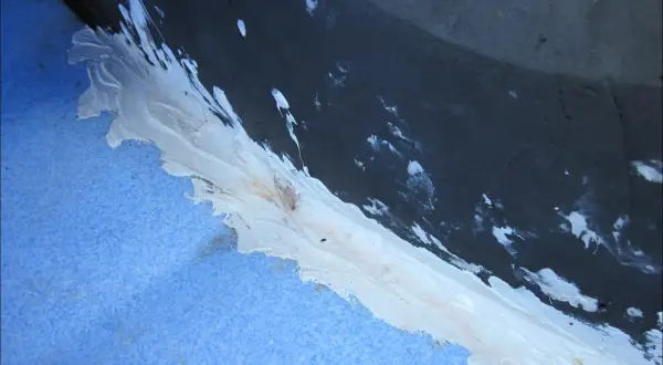Application d'un mastic blanc pour coller du polyéthylène sur une surface bleue texturée, avec des éclaboussures sur le fond sombre.