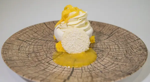 Deliciosa pavlova de mango con merengue, crema batida, coco rallado y salsa de mango sobre un plato rústico.