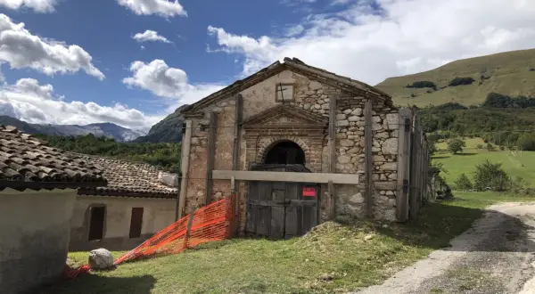 Il rifugio Città di Amandola, con la sua facciata in pietra e legno, è circondato da un paesaggio verdeggiante sotto un cielo azzurro con nuvole bianche.