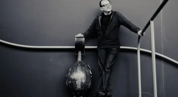 Anouar Brahem, w okularach, opiera się o poręcz, obok niego stoi instrument.