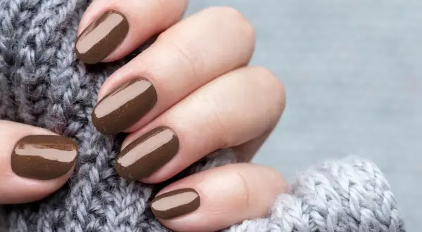 Dłonie z perfekcyjnym manicurem kombinowanym w odcieniu czekolady, otulone miękkim, szarym swetrem.