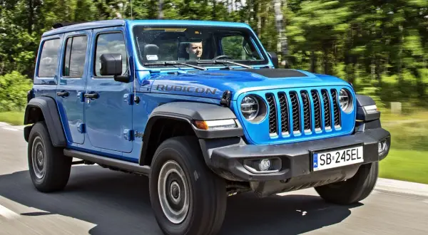 Niebieski Jeep Wrangler 4xe Rubicon z kierowcą za kierownicą jedzie po drodze wśród drzew.