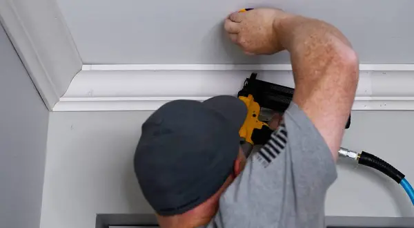 A man is performing crown molding installation, using a nail gun to secure the decorative trim to the ceiling.