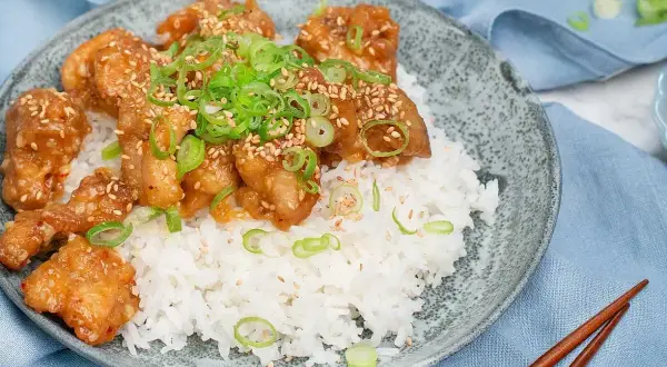 Krispig orange chicken med sesamfrön och salladslök serveras på en bädd av vitt ris. En läcker asiatisk rätt.