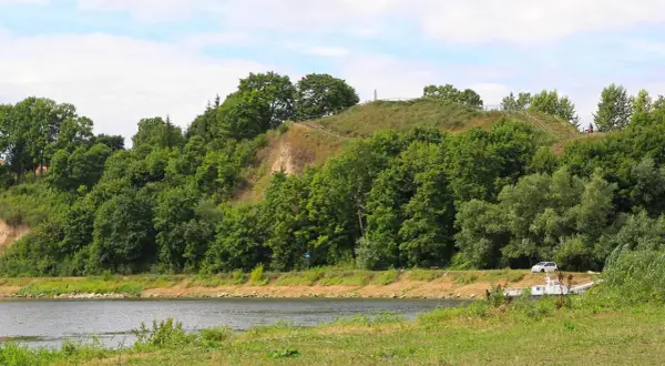 Zielona skarpa nad rzeką, na szczycie której widać ścieżki i ogrodzenie. To malownicza góra zamkowa w Drohiczynie.