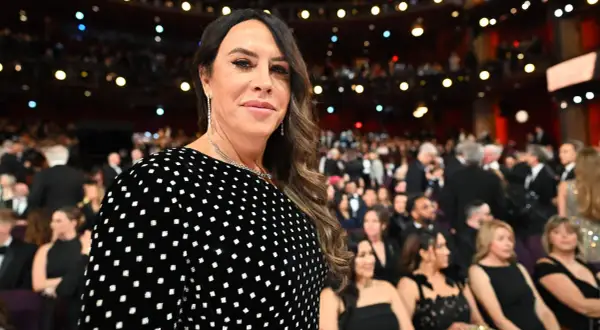 Karla Sofía Gascón, con un vestido negro de lunares blancos, sonríe en un evento con público al fondo.