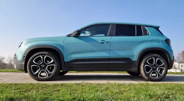 Turkusowy Jeep Avenger, marka samochodu na wprost, stoi na tle błękitnego nieba i zielonej trawy.