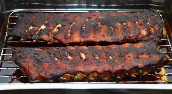Zwei Rippchen-Stränge, dunkelbraun und saftig, ruhen auf einem Rost in einer Bratpfanne. Sie sind perfekt gegart, wie lange Spareribs im Ofen sein sollten.