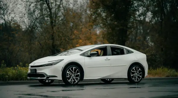 Biała Toyota Prius zaparkowana na mokrej drodze, z drzewami w tle.