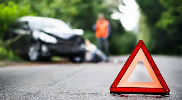 Triangle de signalisation sur la route, près d'une voiture accidentée où une personne gît au sol. Un homme en gilet orange se tient debout. Scène évoquant un homicide involontaire.