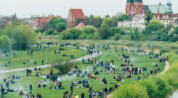 Ludzie odpoczywają na trawie w nadwarciańskiej części Poznania. W tle widać zabytkowe budynki i rzekę.