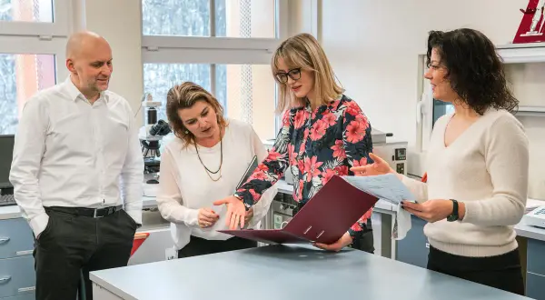 Zespół dyskutuje nad dokumentami w laboratorium. Mężczyzna w białej koszuli, dwie kobiety w bluzkach i jedna w swetrze. W tle mikroskop.