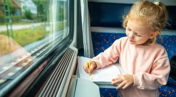Mała dziewczynka rysuje kredką na kartce papieru, siedząc przy oknie w pociągu. Słońce oświetla jej twarz, tworząc magiczny pociąg słoneczny.