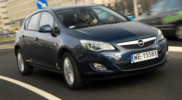 Ciemnoniebieski Opel Astra J 1.7 CDTI porusza się po miejskiej drodze. Kierowca w czapce prowadzi auto.
