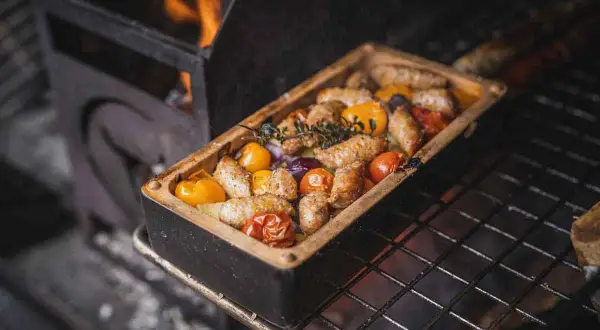 Salsiccia mit Gemüse in einer Auflaufform auf dem Grill, Flammen im Hintergrund.